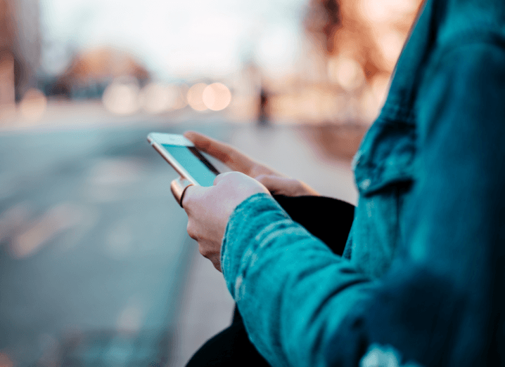 An individual in a blue denim jacket scrolls through an iPhone on an empty street.