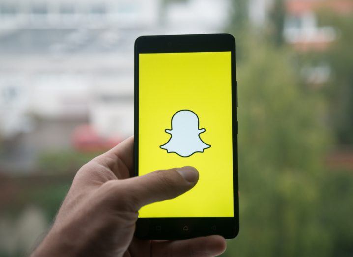 close-up of bright yellow Snapchat logo on smartphone, held up by a hand against a blurry outdoor background.