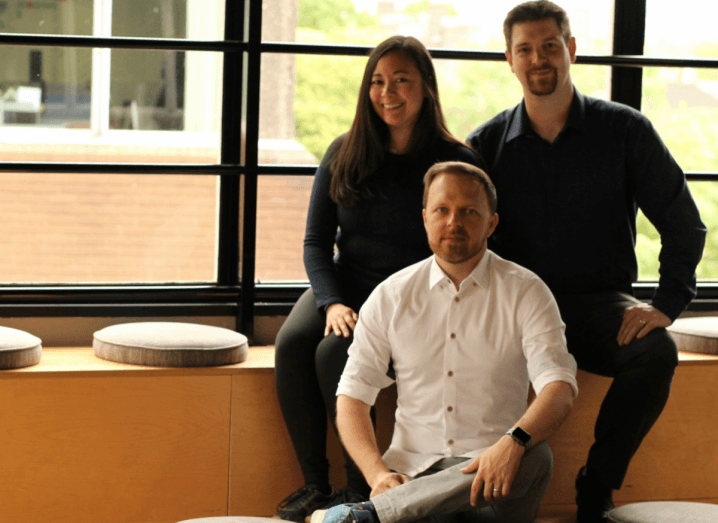 Vela Games' three co-founders sit together on a set of wooden steps. A man and woman wear black, while another man wears a white shirt.