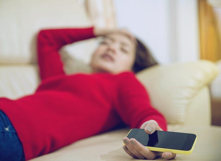 A woman is asleep on a couch with her hand holding her smartphone dangling over the edge.