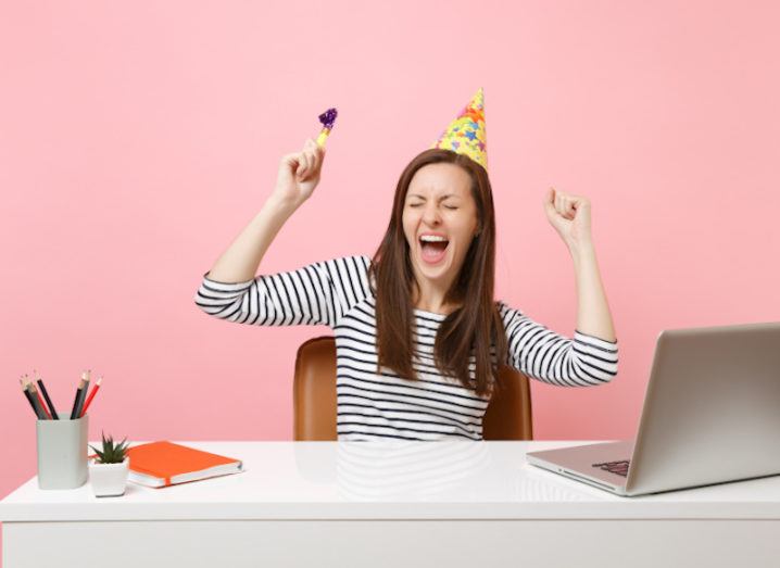 Cheerful girl with closed eyes in birthday party hat with playing pipe screaming celebrating while sit work at desk with pc laptop isolated on pink background.