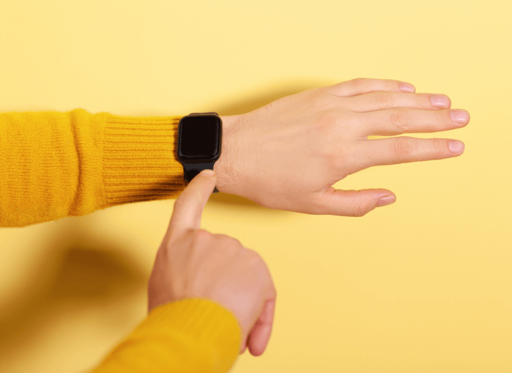 A person wearing a yellow jumper points at an Apple Watch on their wrist, in front of a yellow background.