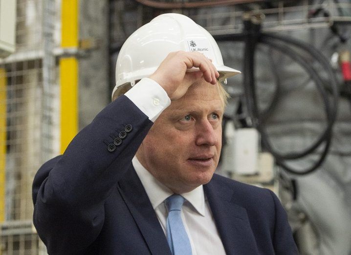 Boris Johnson wearing a navy suit and blue tie places a white hard hat on his head while visiting an energy research centre.