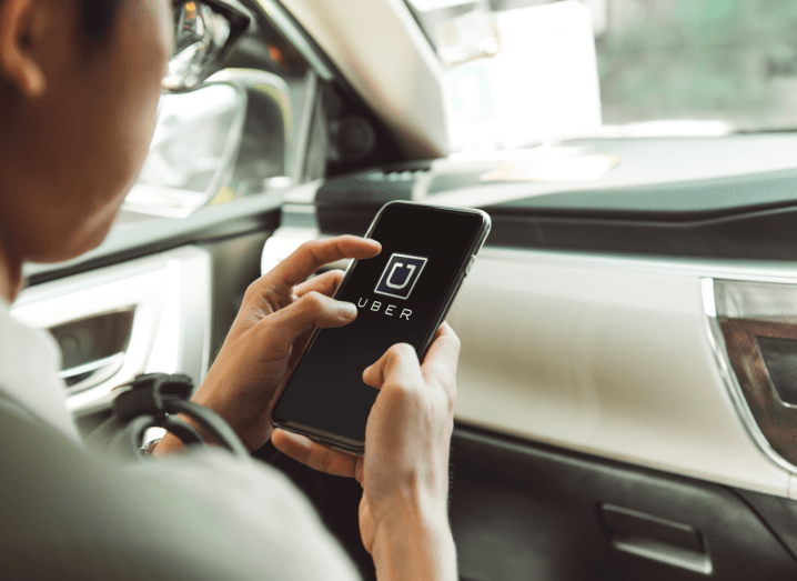 A man sitting in a car looking at his mobile phone, which has the Uber app open onscreen.