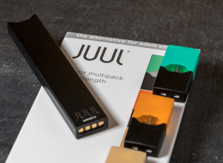 A black plastic Juul e-cigarette, which resembles a USB stick sitting on top of a white box that reads Juul. Beside the e-cigarette, there are colourful refill pods.