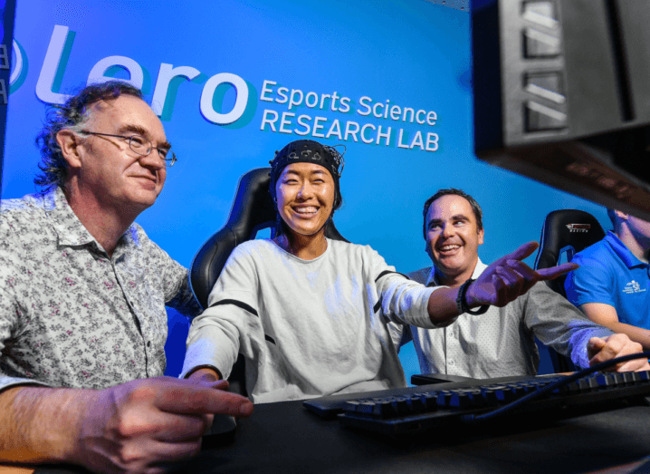 Three people sit in front of a computer. The person in the middle is wearing headgear connected to the computer. There is a blue wall in the background that says 'Lero' on it.