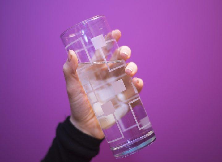 A hand holding a glass of water against a purple background.