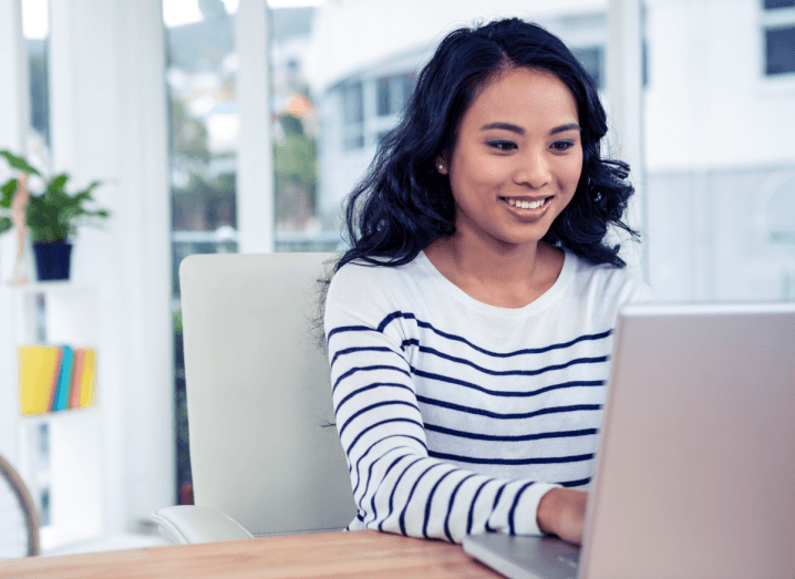 A woman with dark hair wearing a white long-sleeved shirt with navy stripes smiles in an office as she uses a laptop.