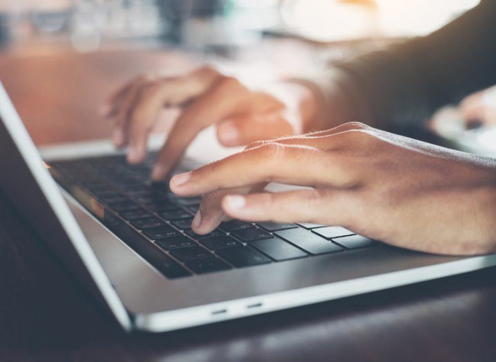 View of hands typing on a silver laptop keyboard with black keys on a wooden table.