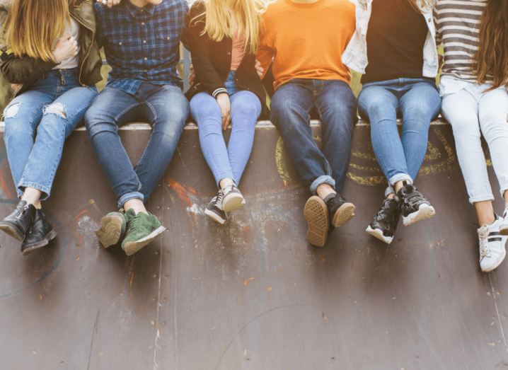 A group of teenagers sitting on a skateboard ramp wearing jeans and trainers. They are all wearing colourful shirts and sweaters.