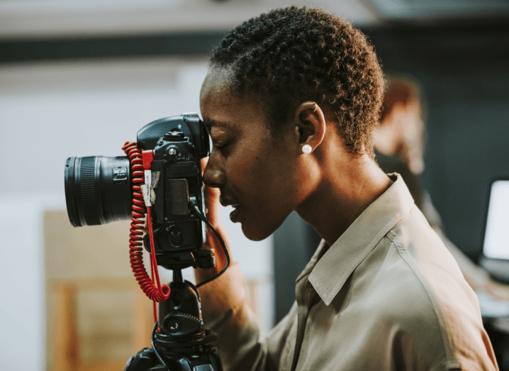 A woman in a beige mac jacket closes one eye as she looks into a camera's viewfinder.