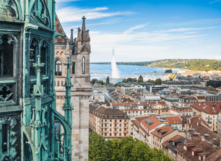 A view of Geneva from the Cathedral of Saint-Pierre. There are buildings with red-tiled roofs in front of a lake with a large fountain in it.