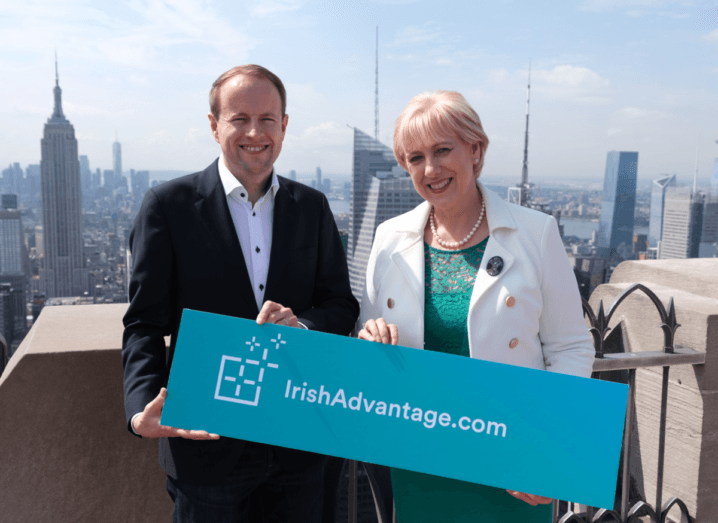 A man in a black suit and white shirt stands on top of the Rockefeller building with a woman in a green dress and a white jacket, holding a blue sign that reads Irish Advantage.