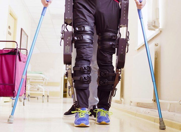 Person wearing an exoskeleton using crutches to walk down a hallway.