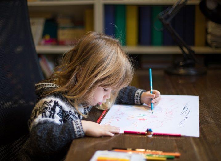 Blonde child in a grey sweater sitting and painting a white sheet paper with a blue pencil using their left hand.