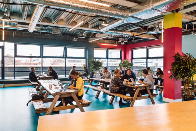 A room with windows along every wall and men and women eating food at picnic benches within it