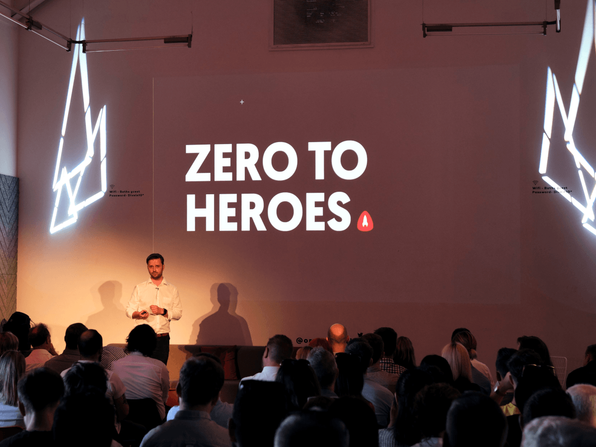A man stands beside a projector screen that reads "Zero to heroes" in front of an audience in a dark room. He is wearing a white shirt and black trousers and has his hands folded in front of his body.
