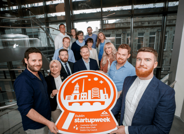 Fourteen entrepreneurs stand at the bottom of a large spiral staircase in a glass office building holding a large orange sign depicting Dublin in a snowglobe. The sign reads "Dublin Startup Week".