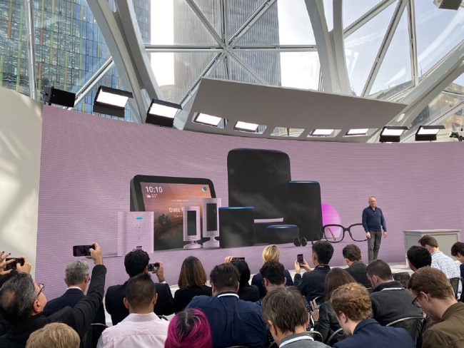 A man speaks to an audience gathered at Amazon’s Seattle headquarters in front of a backdrop displaying an array of devices.