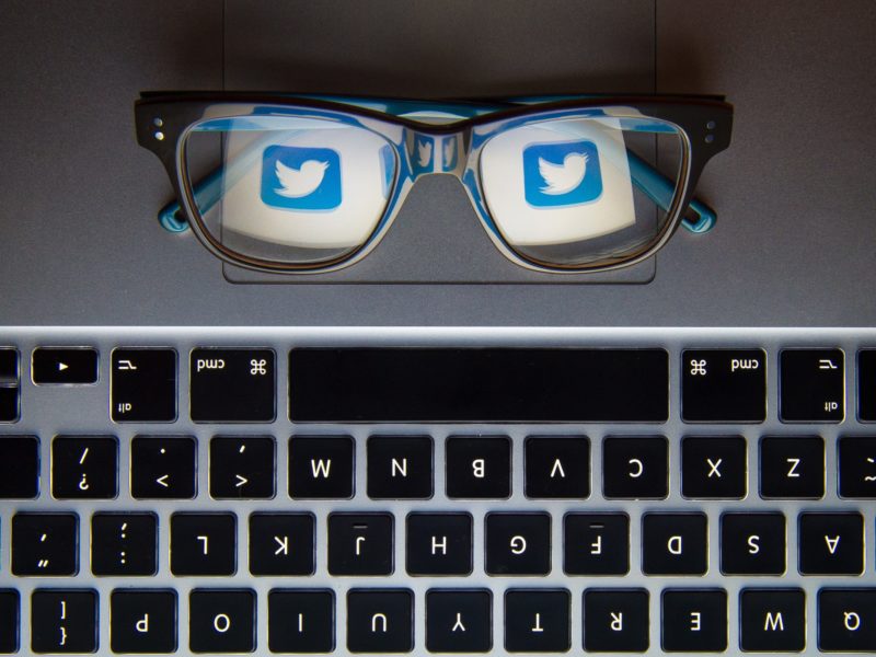 Pair of glasses sitting on a desk beside a keyboard, with twitter logo reflected in the glass.