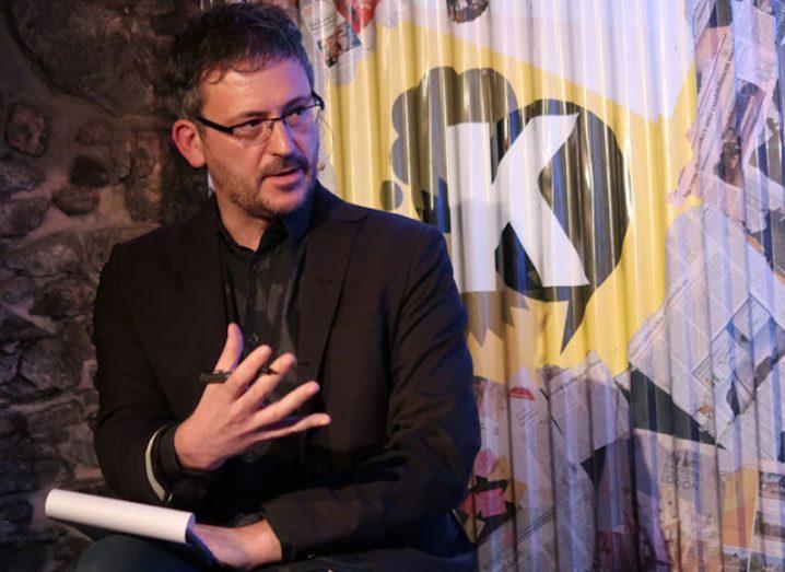John Cleere, a man in a black suit and shirt, wearing glasses, sits with a notepad in front of a colourful wall on which is a graphic letter K.