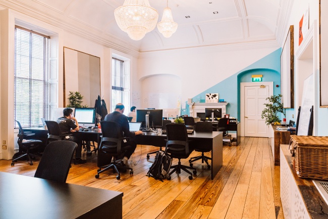 A bright office space with men and women working at computers at desks, with a door on the back wall closed. 