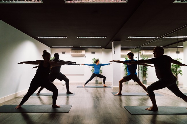 Men and women are pictured on yoga mats in a room with a wooden floor, copying a pose from a yoga instructor