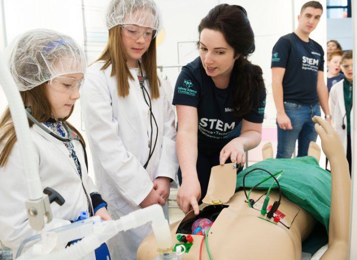 Two girls wearing lab coats, hair nets and stethoscopes are shown the heart inside a human surgery dummy by a festival volunteer, as another volunteer waits by a queue of children looking on in wonder.