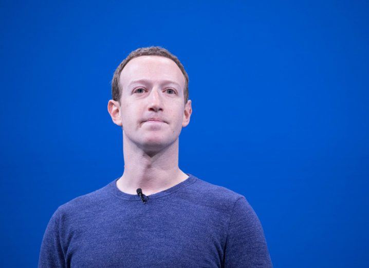 View of Mark Zuckerberg in blue sweater standing against blue background.