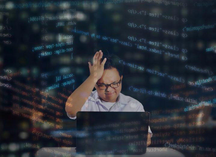 Young male programmer with glasses getting stressed out while coding. Shot behind office window at night with HTML codes surrounding the programmer.