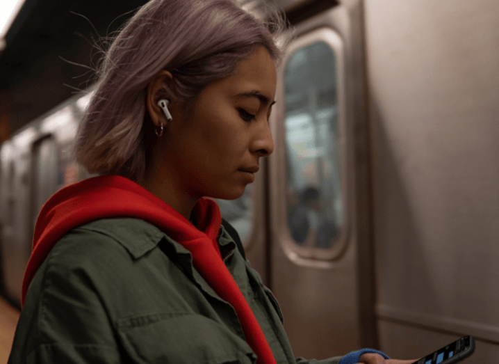 A woman with lilac hair stands in front of a subway train passing by, wearing an orange hoodie and a green jacket over it. She has the white AirPods Pro in her ear.