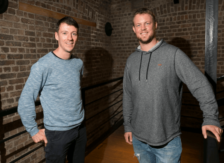 A man in a blue jumper standing with his hands in the pockets of his black jeans, who has black hair, standing beside another man in a grey hoodie and blue jeans. The man on the right has auburn hair. They are standing in a stairwell in front of an exposed brick wall.