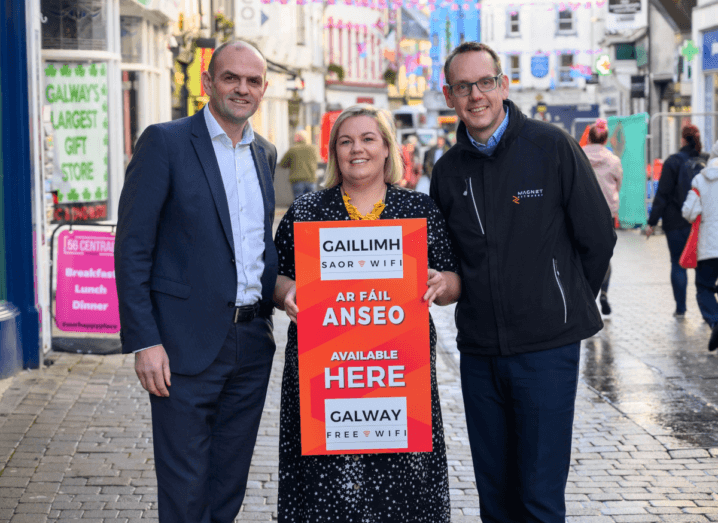 A man in a navy suit and blue shirt stands on the cobblestone streets of Galway beside a woman in a black dress who is holding a red sign that says "Galway Free Wi-Fi Available Here". To the right of that woman is a man in a navy coat and blue shirt. He has glasses and dark hair. The woman has blonde hair and the man on the left of her is bald.