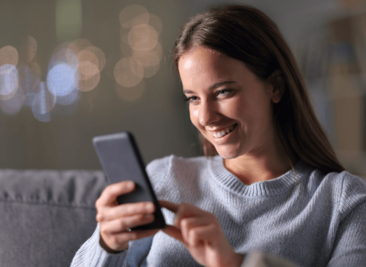 A woman in a light blue jumper sits on a grey sofa playing a game on a black mobile phone. She has long, brown hair and is smiling.