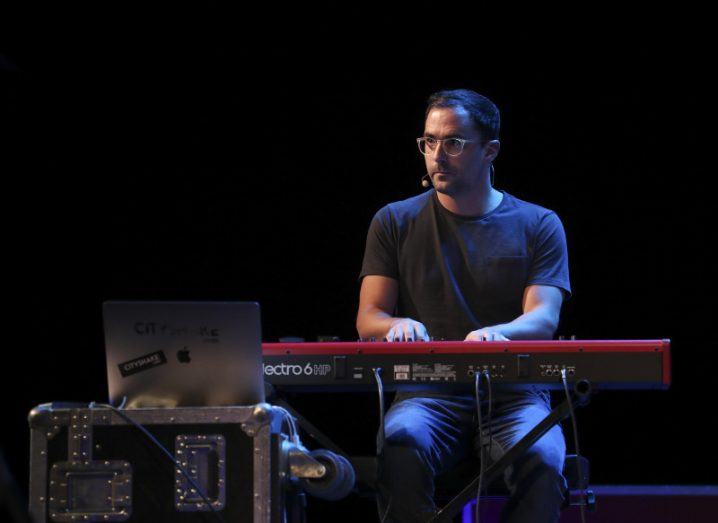 View of man on stage with glasses sitting at keyboard piano playing against black background.