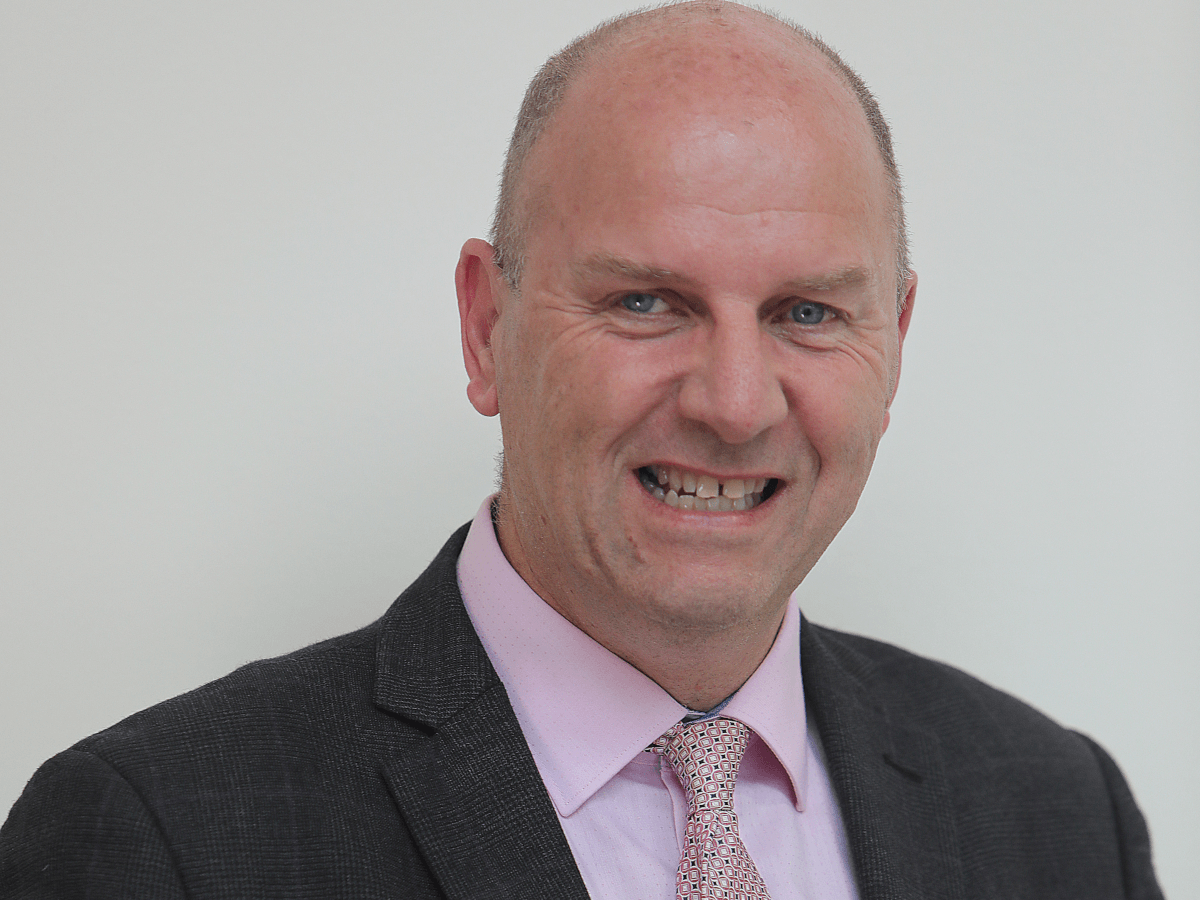 A bald man smiling in a pink shirt, pink tie and grey suit. 