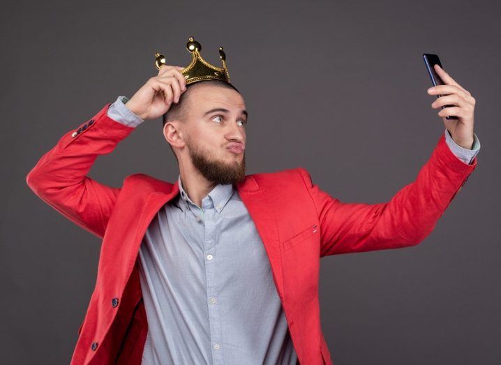 Young, bearded man in gold crown taking a selfie.