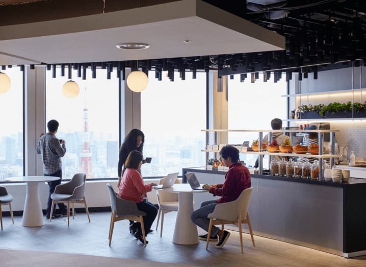 The staff kitchen in Google's Tokyo office, overlooking the Tokyo Tower in the Shiba-koen district of Minato. The view of Tokyo's skyline is visible outside of the window, where an employee is leaning against a pillar gazing outside. There are two employees sitting at a round table using laptops while another employee stands beside the table chatting to them. Behind them, there is a bar full of breakfast cereals and snacks, as well as plates and bowls. There is an employee standing beyond the cereal selection, presumably using a microwave.
