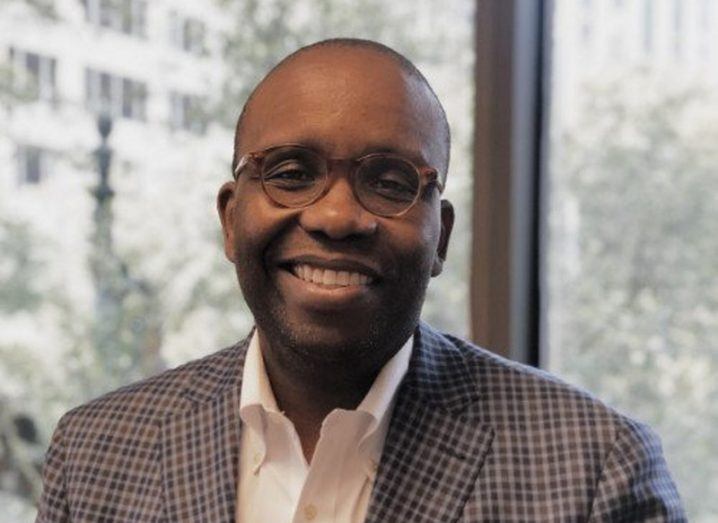 Headshot of a smiling man wearing a check blazer, white shirt and tortoiseshell glasses, pictured against a window overlooking an office courtyard.