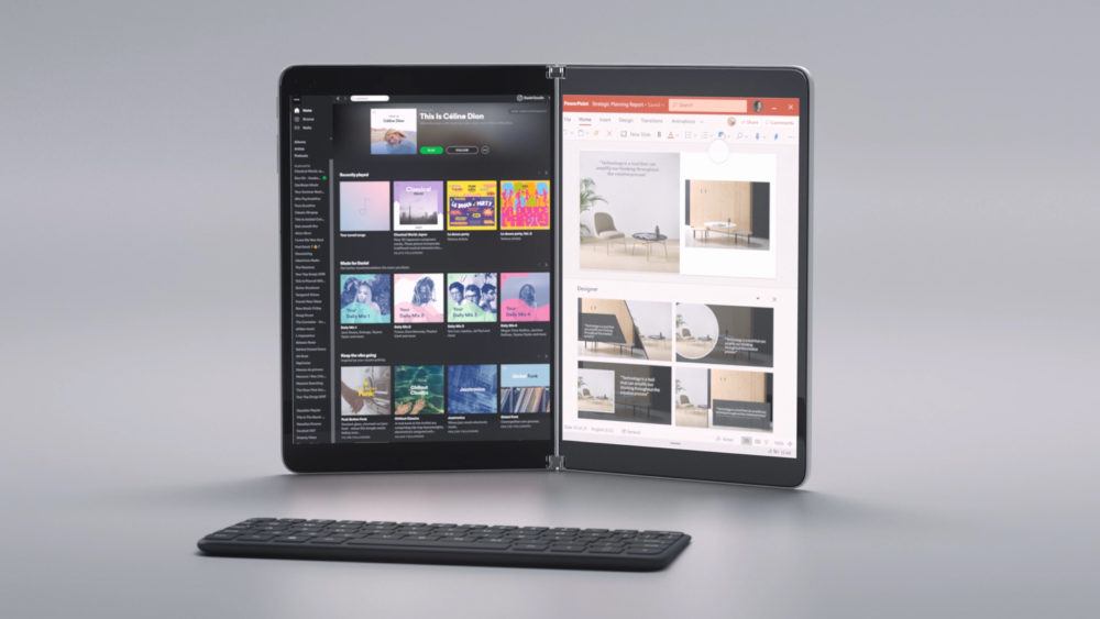 A foldable, dual-screen tablet which is standing up on its hinge in front of a keyboard that can be used with it.