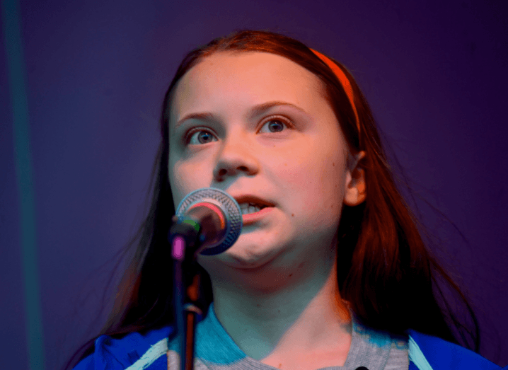 A young woman speaking into a microphone. She has clear skin and brown hair, which is held back by a red hairband. The photograph is taken from the shoulders up, so her purple hoodie is just about visible in the shot. She is standing in front of a purple background.