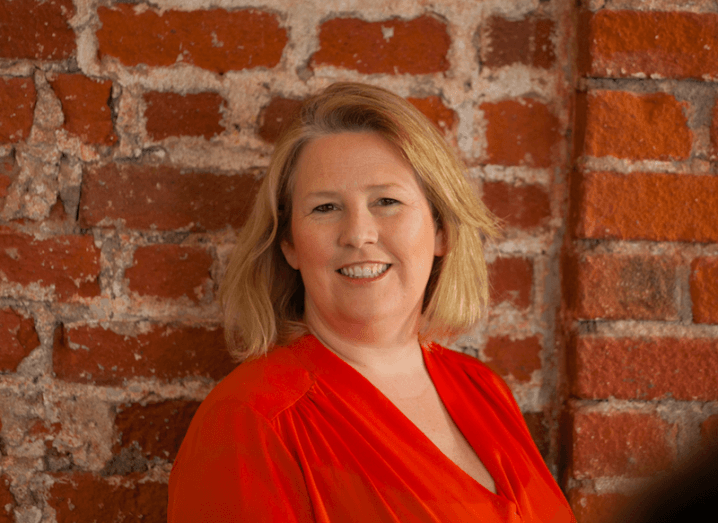 A woman with blonde hair and a red shirt stands against a red-brick wall.