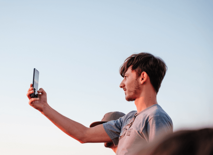 A man in a grey T-shirt stands in front of a blue sky, holding his phone out with one arm to take a photograph.