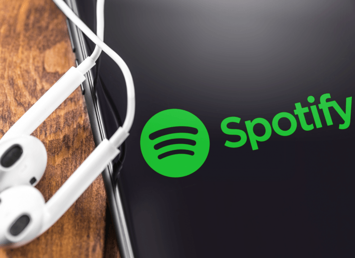 An iPhone screen displaying the Spotify logo. The phone is on a wooden surface with a pair of Apple AirPods beside it.