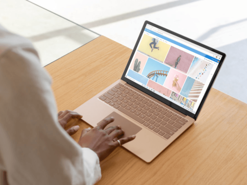 A woman with her hand on the touchpad of the Surface Laptop 3. The laptop is on a light-coloured wooden table. The device appears to be rose gold or gold.