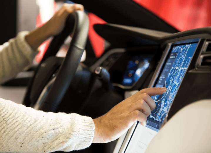 A person in a beige knitted jumper sitting behind the steering wheel of a Tesla, touching the computer built into the car's console.