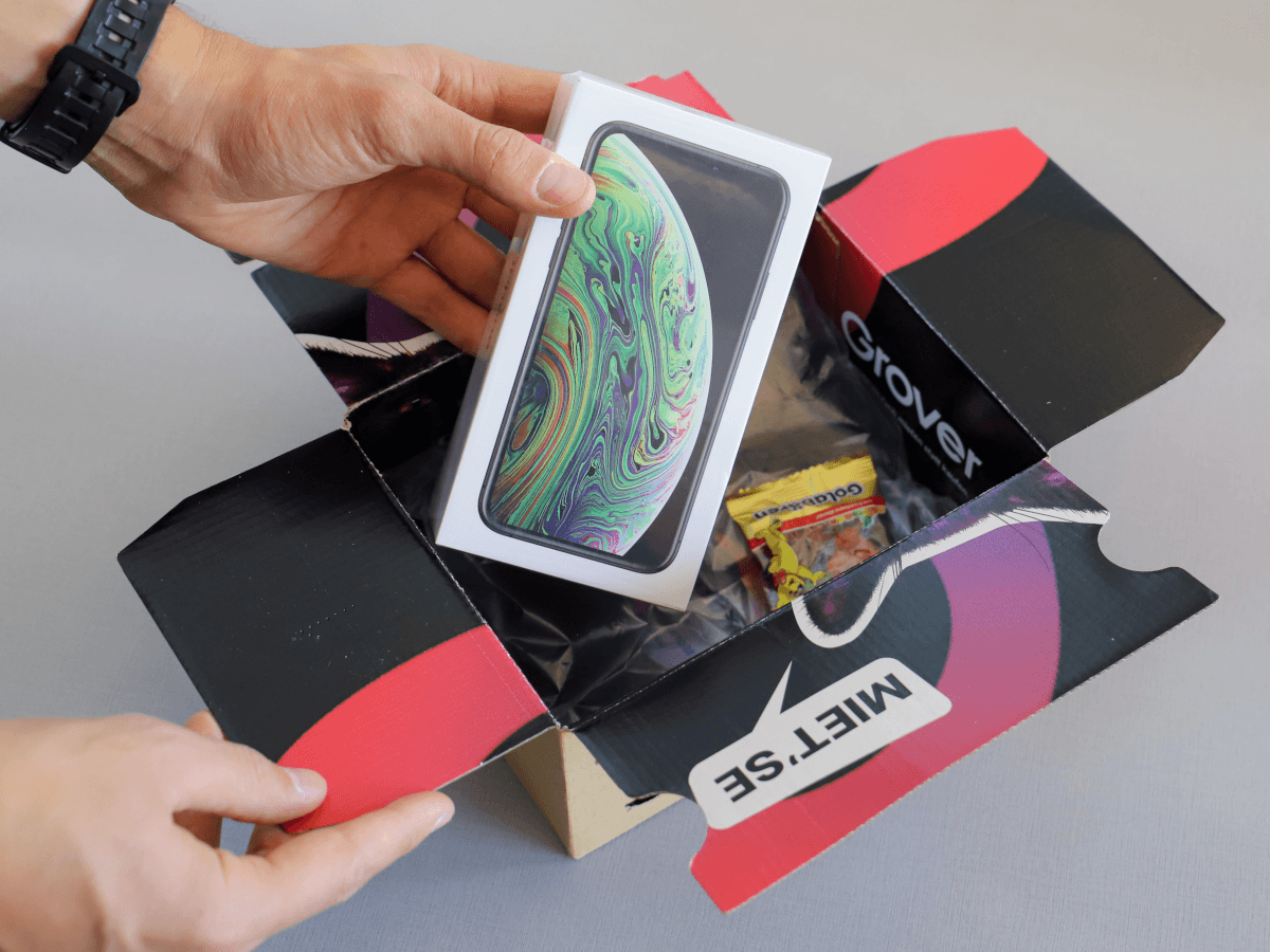 A hand holding an iPhone XR in its packaging beside a colourful box it was presumably delivered in.