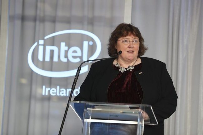 A woman in a burgundy velvet top and black jacket addresses an audience from a Perspex podium in front of a lit-up Intel logo.