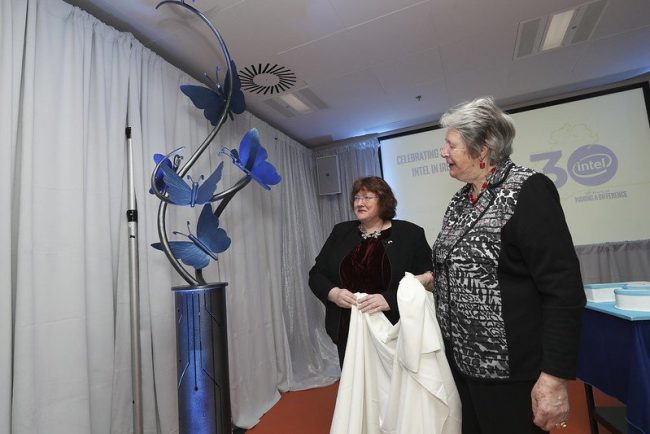 Two women hold a cotton sheet after pulling it away to reveal a large blue ironwork sculpture of butterflies emanating from a pillar of electronic connectors.