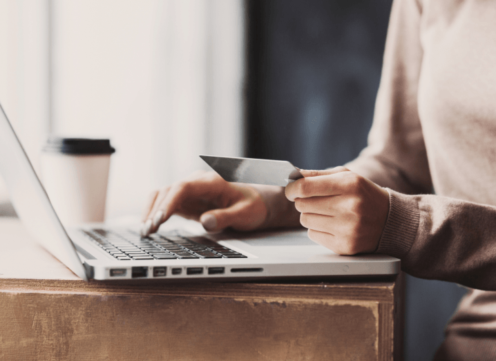 A person typing on a MacBook with one hand and holding a payment card in the other hand. They have a coffee on the the table beside them in a disposable cup. They are wearing a beige jumper and grey nail polish.